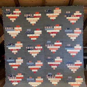 May include: A large rectangular quilt featuring a dark gray background with a repeating pattern of sixteen heart shapes. Each heart is constructed from horizontal fabric strips in various floral, plaid, and solid patterns of red, blue, and white.