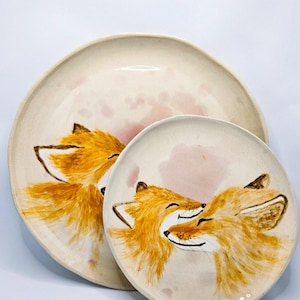 Peut inclure: Deux assiettes en céramique avec des illustrations de renards peintes à la main. Les renards sont orange et marron, avec un fond rose. Les assiettes ont une base crème et une forme irrégulière. Mesures non disponibles.