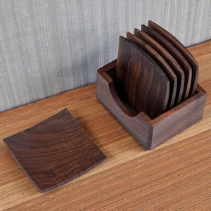 Op de afbeelding: Een set van zes donkerbruine houten onderzetters met een gebogen ontwerp, gepresenteerd in een bijpassende houten houder. Eén onderzetter is apart, met de houtnerf zichtbaar. De onderzetters liggen op een houten oppervlak.