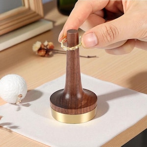 以下が含まれることがあります： 濃い茶色の仕上げと金色のベースを備えた木製のリングディスプレイスタンド。 手は、スタンドにテクスチャデザインのゴールドリングを持っています。 スタンドは白い布の上にあります。