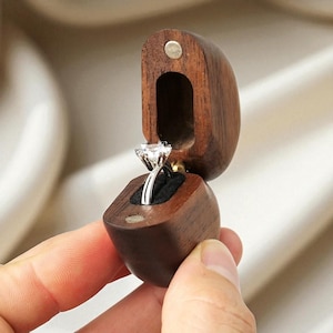 Può includere: Un anello di fidanzamento con diamanti presentato in un portagioie di legno marrone scuro aperto. L'anello presenta un grande diamante rotondo trasparente su una fascia d'argento. Il portagioie è aperto, rivelando l'anello contro un interno in velluto nero.