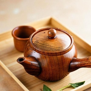 May include: A handcrafted wooden teapot with a matching lid and a small cup, all in a warm brown hue. The teapot features a curved handle and spout, set on a light-colored wooden tray.