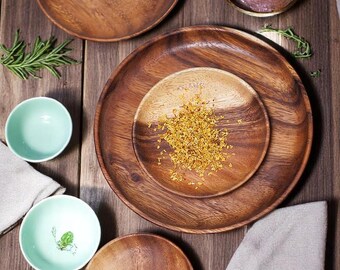 May include: Overhead shot of wooden plates and bowls on a wooden surface. The plates vary in size and shape, with a large round plate in the center. Small, light green bowls and a sprig of rosemary are also present.