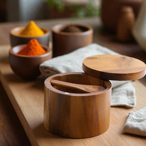 Könnte beinhalten: Gewürzbehälter aus Holz mit Deckeln und kleinen Löffeln. Die Behälter sind mit farbenfrohen Gewürzen gefüllt, darunter orangefarbene, gelbe und braune Pulver. Die Behälter stehen auf einem Holzschneidebrett mit einem Leinentuch.