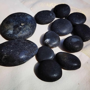 May include: A collection of smooth, dark gray river stones of varying sizes. The stones are arranged on a light-colored, textured surface. Some stones have subtle color variations and natural markings.