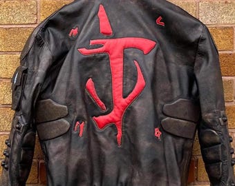May include: A brown leather jacket with a red embroidered symbol on the back. The symbol is a stylized 'T' with a downward facing hook on each side. The jacket has a zipper closure and two pockets on the front.