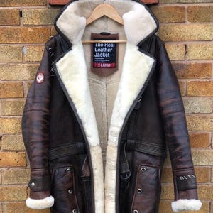 May include: A brown leather shearling lined jacket with a fur collar. The jacket has a belt, two pockets, and a label that reads "The Real Leather Jacket" and "X-Large".