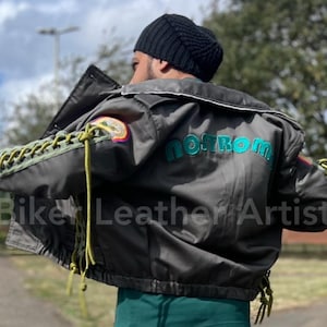 May include: A dark grey bomber jacket featuring the word "nostrom" in teal on the back. The jacket has white trim, patches on the sleeves, and olive green lacing details. The sleeves are laced with olive green cords. The text "Bike Leather Artist" is at the bottom.