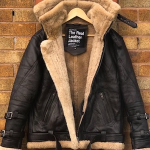 May include: A brown leather aviator jacket with a shearling lining and a fur collar. The jacket has a zipper closure, two front pockets, and a label that reads "The Real Leather Jacket".