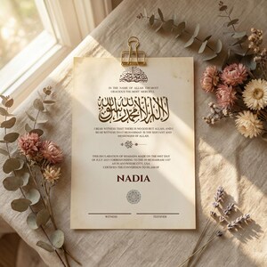 May include: A beige certificate featuring Arabic calligraphy and English text, secured by a gold clip. The certificate includes the name "Nadia" and is decorated with floral arrangements. The text states the declaration of Shahada.