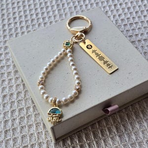 May include: A gold-colored keychain with a pearl and gemstone accent. The keychain features a Spotify code tag and a charm with a blue and gold design. The keychain is resting on a light gray box.