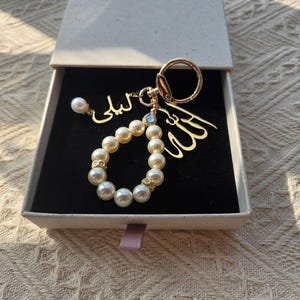 Puede incluir: Un llavero dorado con un rosario de perlas, un colgante de perlas y caligrafía árabe. El llavero se presenta en una caja de regalo blanca con interior negro. El rosario está hecho de perlas blancas grandes y redondas.