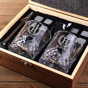 May include: A wooden gift box containing two engraved whiskey glasses, a metal ice spoon, and ice cubes. The glasses are etched with the letter "H" inside a laurel wreath and the name "Hunter". The box is lined with black fabric.