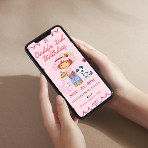May include: A smartphone displays a pink birthday invitation. The invitation features a cartoon illustration of a girl with a strawberry hat, holding a cake, with a cat and a dog. The text reads "Cindy's 2nd Birthday" and includes date, time, and RSVP information.