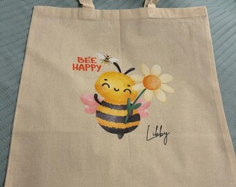 May include: A beige tote bag with a bee design. The bee is holding a daisy and has the words "Bee Happy" above it. The name "Libby" is written below the bee.