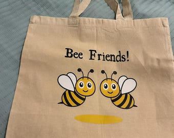May include: A beige canvas tote bag with black handles and the text "Bee Friends!" printed on the front. Two cartoon bees with black and yellow stripes are flying above a yellow circle.