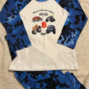 May include: White long-sleeve shirt with blue camouflage sleeves. The shirt has a graphic of monster trucks and the text "When I wake up I will be" and "Ted".
