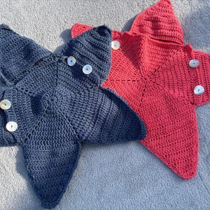 May include: Two star-shaped crochet blankets, one in dark blue and one in red. Each blanket features a hood at the top point and is accented with white circular buttons along the side edges, showcasing a textured, hand-stitched yarn construction.