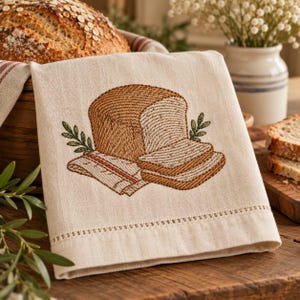 May include: A cream-colored linen kitchen towel featuring an embroidered design of a loaf of bread and two slices, accented with green sprigs. The towel has a decorative hemstitch detail along the bottom edge and a soft, woven texture.