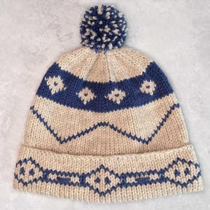Könnte beinhalten: Eine beige Strickmütze mit einem blauen geometrischen Muster und einem weißen Bommel oben.