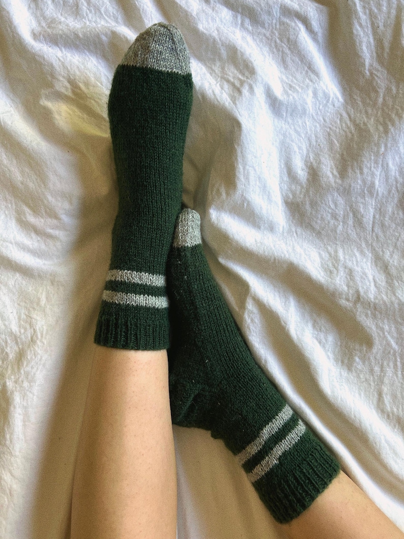 May include: A pair of green socks with white stripes. The socks are made of a soft, knitted fabric and have a grey toe and heel.