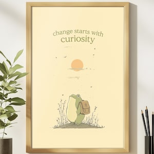 May include: A framed art print with a cartoon frog wearing a backpack, set against a light yellow backdrop with a sun and the words "change starts with curiosity". The frog stands on a small hill. The frame is made of light brown wood.