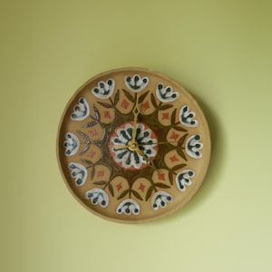 May include: A round, decorative wall clock with a floral design. The clock face is tan with a central floral pattern in red, black, and white. The clock hands are gold. The clock is mounted on a light green wall.