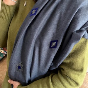 May include: A blue scarf with diamond-shaped embellishments. The scarf has a soft texture and is draped over a green sweater. The diamond shapes are a darker blue with a pattern of small, silver-colored studs.