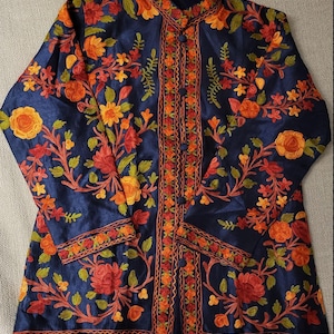 Pode incluir: Uma jaqueta de seda azul marinho com gola mandarim e fecho de botões. A jaqueta é bordada com desenhos florais coloridos em tons de laranja, vermelho, amarelo e verde.