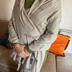 May include: A beige scarf with a geometric pattern of dark brown rectangles. The scarf is draped over a green sweater. The scarf appears to be made of a soft, lightweight material. The scarf is a neutral color and could be worn with many outfits.