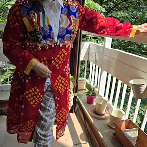 Op de afbeelding: Een rood jasje met patronen, kleurrijke patchwork en gouden randen. Het jasje wordt gedragen over een wit overhemd met kraag en gecombineerd met een geruite broek. Het jasje heeft een uniek ontwerp met diverse patronen en versieringen.