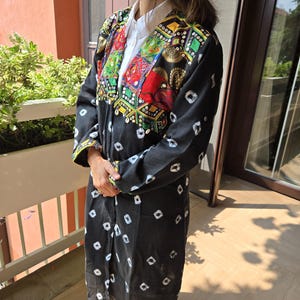 Puede incluir: Chaqueta negra de manga larga con patrones blancos en forma de diamante. La chaqueta presenta detalles bordados coloridos en el cuello y la parte superior del pecho, incluyendo acentos rojos, verdes y dorados. Se lleva sobre una camisa blanca.