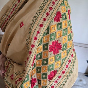 Peut inclure: Châle beige avec broderies complexes. Le motif comprend des motifs floraux rouges, des motifs géométriques verts et jaunes, et des coutures décoratives sur les bords. Le châle est drapé, mettant en valeur le savoir-faire.