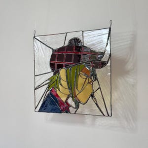 May include: A stained glass window depicting a person with green hair wearing a red and black plaid hat. The person is looking to the right. The stained glass is framed in black metal.