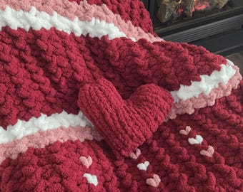 May include: A handmade, chunky knit blanket in a deep red color, accented with stripes of white and light pink. The blanket features small heart-shaped embellishments and a matching heart-shaped pillow. The cozy blanket is perfect for snuggling.