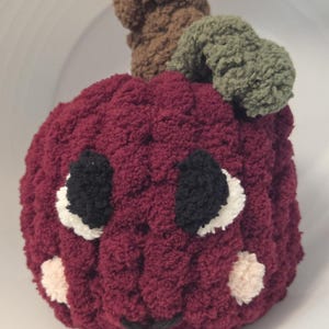 May include: A plush, burgundy-colored apple-shaped toy with black and white eyes, a black smile, and two small pink cheek accents. The apple has a brown stem and a green leaf. The toy is made of a soft, textured material.