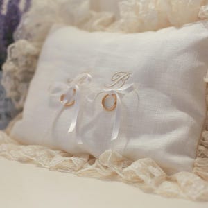 May include: A white linen ring pillow with gold wedding bands tied with white ribbons. The pillow is embroidered with a gold initial and is trimmed with lace. A small pot of lavender is in the background.