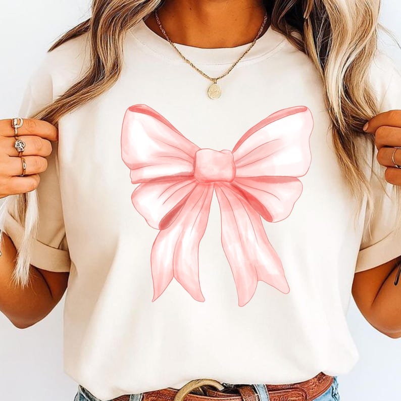 May include: A cream-colored short-sleeve t-shirt featuring a large, centered graphic of a soft pink ribbon bow. The bow design has detailed shading and folds, creating a dimensional appearance on the front of the garment.
