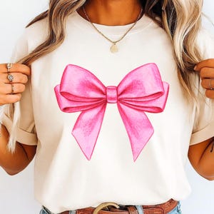 May include: A cream-colored short-sleeve t-shirt featuring a large, centered graphic of a bright pink bow. The bow design has a hand-drawn appearance with visible shading and texture. The t-shirt has a crew neckline and a relaxed, casual fit.