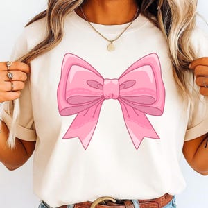May include: A cream-colored short-sleeve t-shirt featuring a large, centered graphic of a pink bow. The bow is illustrated with various shades of pink to create depth and highlights, showcasing a classic ribbon shape with two loops and two tails.