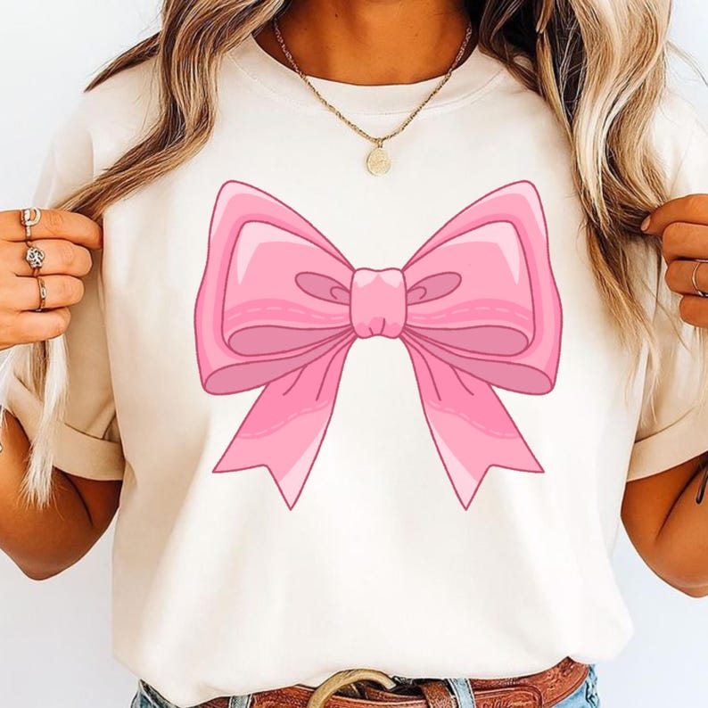 May include: A cream-colored short-sleeve t-shirt featuring a large, centered graphic of a pink bow. The bow is illustrated with various shades of pink to create depth and highlights, showcasing a classic ribbon shape with two loops and two tails.