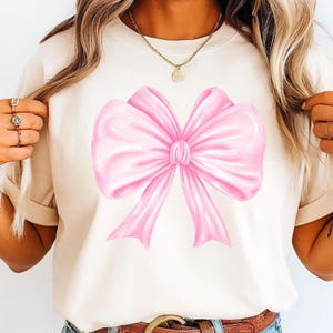 May include: A cream-colored short-sleeve t-shirt featuring a large, centered graphic of a glossy, light pink ribbon bow. The bow has soft, rounded folds and two downward-pointing tails, creating a smooth, satin-like appearance on the fabric.
