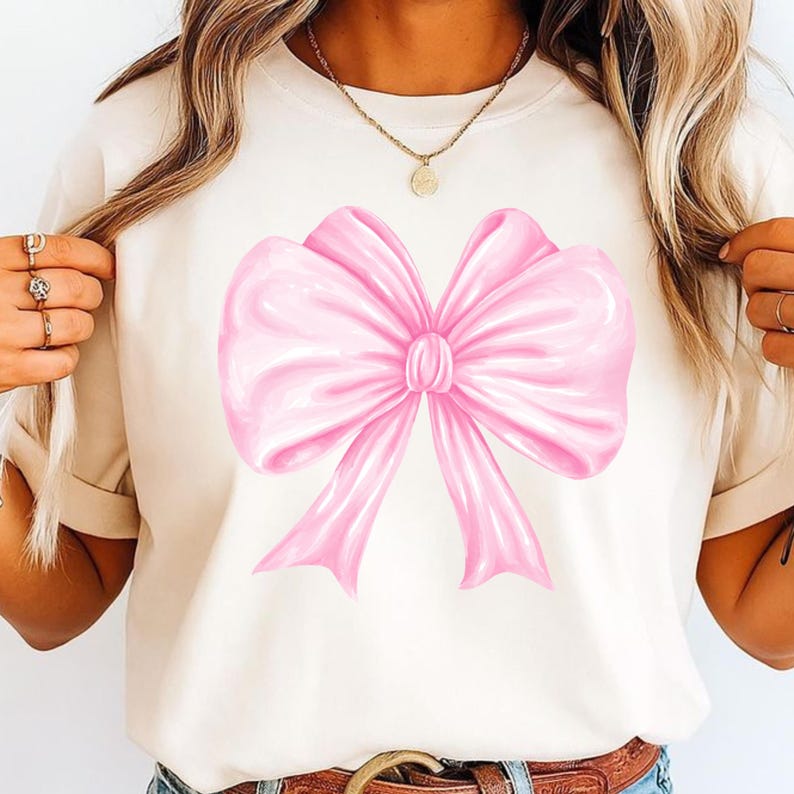 May include: A cream-colored short-sleeve t-shirt featuring a large, centered graphic of a glossy, light pink ribbon bow. The bow has soft, rounded folds and two downward-pointing tails, creating a smooth, satin-like appearance on the fabric.