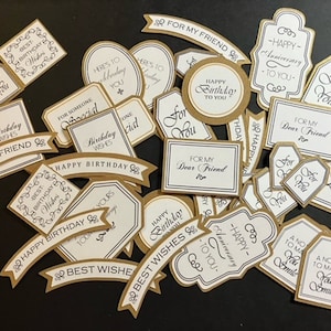 May include: A collection of decorative gift tags in various shapes, including banners, ovals, and rectangles. The tags feature gold borders, white centers, and elegant black script with phrases like "Happy Birthday" and "For You."