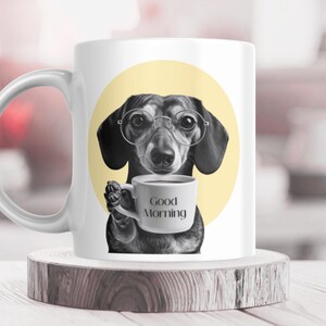 May include: White ceramic mug with a black and white illustration of a dachshund wearing spectacles and holding a coffee cup. The cup has the words "Good Morning" printed on it. The background is a yellow circle.