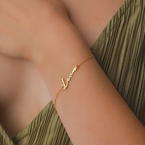 Könnte beinhalten: Ein zartes, goldfarbenes Armband mit dem Namen "Leona" in Schreibschrift. Das Armband wird an einem Arm mit einem grünen Oberteil getragen. Die Kette des Armbands ist dünn und der Name ist der Blickfang.