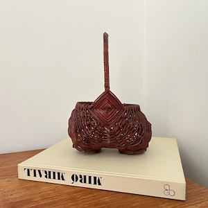Op de afbeelding: Een roodbruine geweven mand met een handvat. De mand heeft een unieke vorm met een ruitvormig ontwerp aan de bovenkant. Het staat op een boek met de titel "MIRÓ MIRALL".