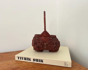 May include: A reddish-brown woven basket with a handle. The basket has a distinctive shape with a diamond design at the top. It rests on a book titled "MIRÓ MIRALL".