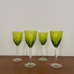 May include: Four green-tinted wine glasses with clear stems and bases are arranged on a wooden surface. The glasses have a decorative, etched pattern on the bowl. The background is a plain white wall.