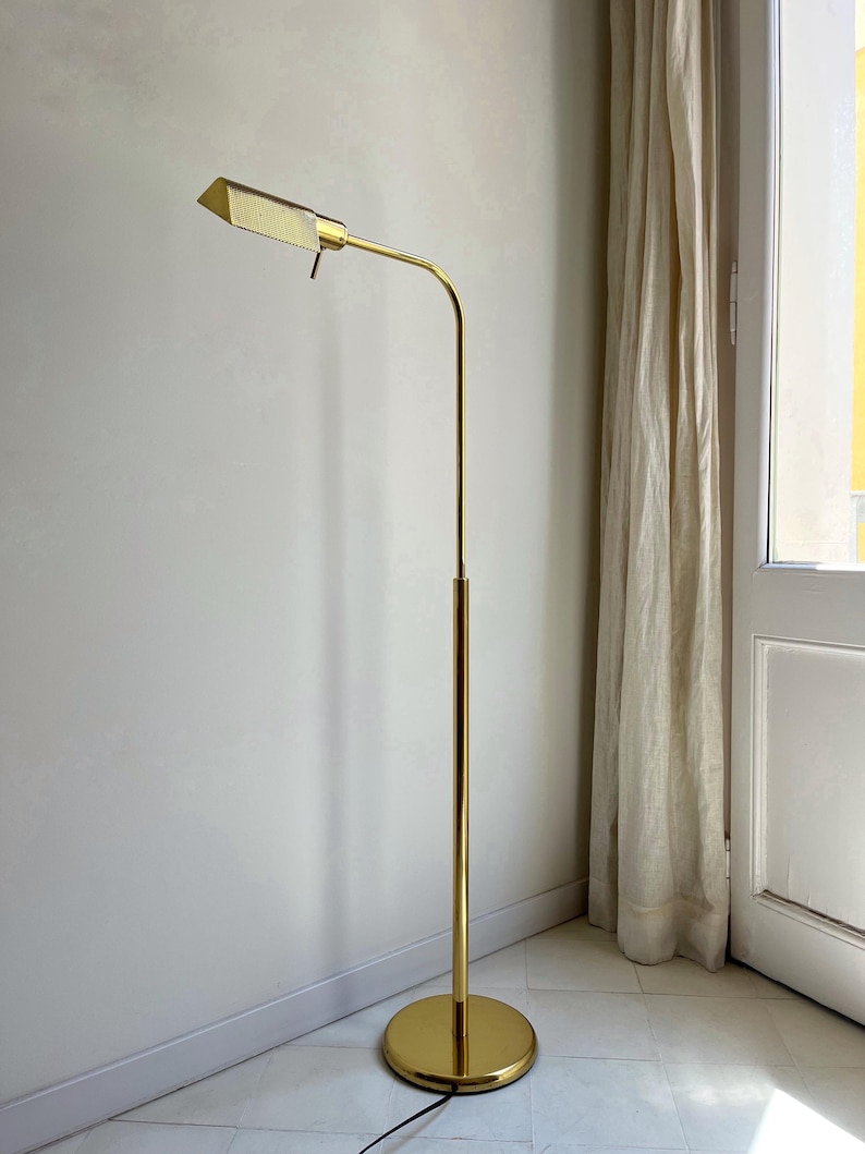 May include: A vintage gold floor lamp with a rectangular head and a curved arm. The lamp has a straight pole and a round base. The lamp is made of metal and has a textured lampshade. The lamp is positioned in a corner near a window with a curtain.